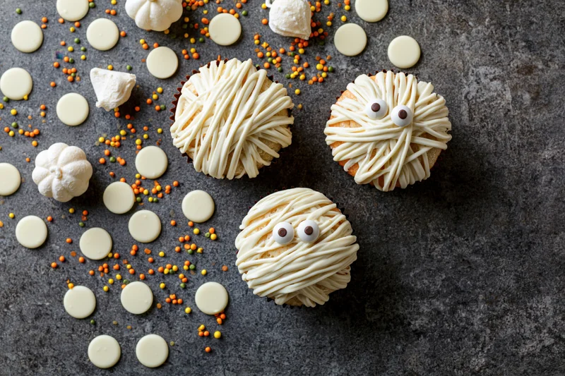 Three halloween mummy cupcakes with white icing drizzle and candy eyes on dark surface surrounded by sprinkles and white chocolate discs