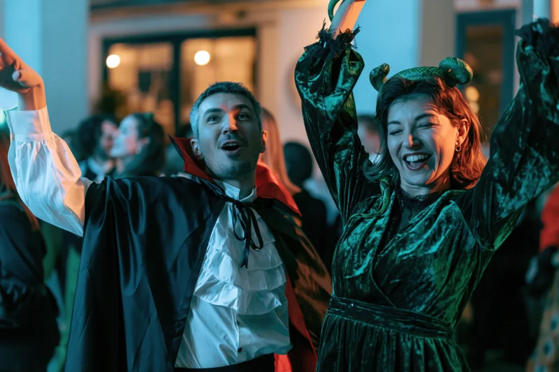 Man in vampire costume and woman in witch costume dancing with arms raised at outdoor halloween party