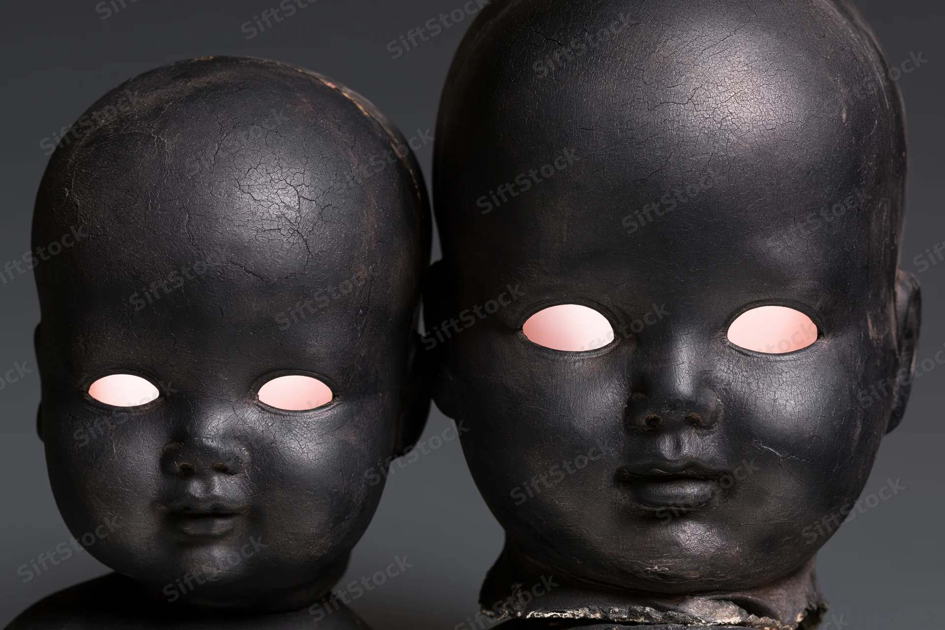 Two black-painted antique doll heads side by side, one smaller, with glowing pink eyes in a dark studio setting