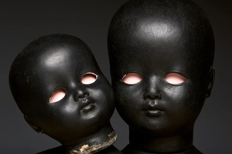 Two black-painted antique doll heads side by side, one smaller, with pink eye sockets, cracked surface, studio lighting