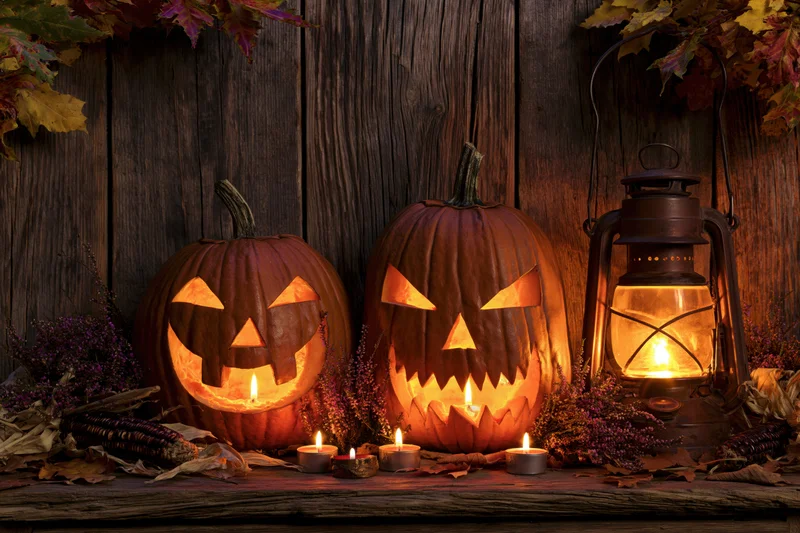 Two carved jack-o-lanterns glowing with candlelight on a wooden table surrounded by autumn foliage and a lantern