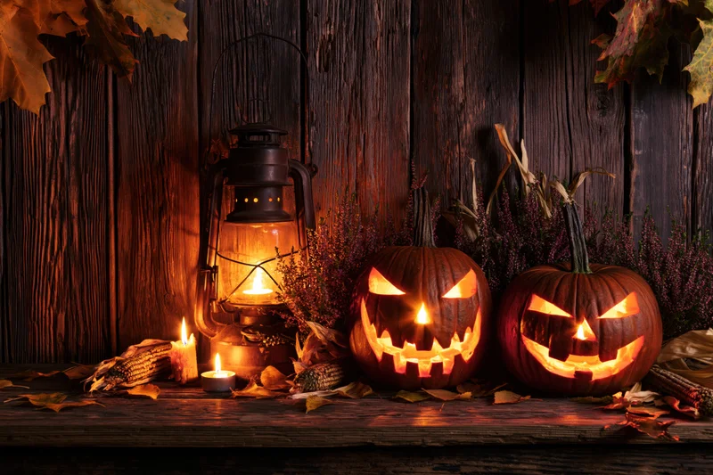 Two carved jack-o-lanterns glowing with candlelight on a rustic wooden table surrounded by autumn leaves and corn husks