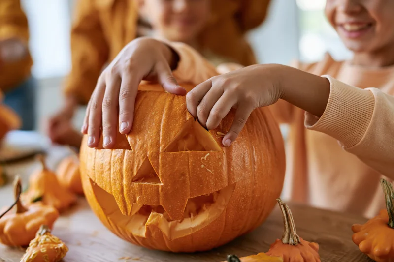 Children's hands carving a jack-o-lantern on a wooden table surrounded by small pumpkins