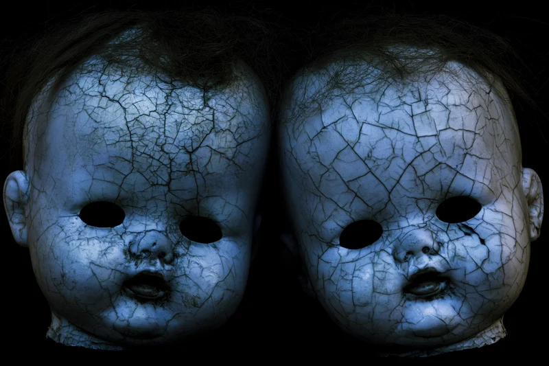 Two cracked vintage baby doll heads side by side with hollow black eyes against dark background