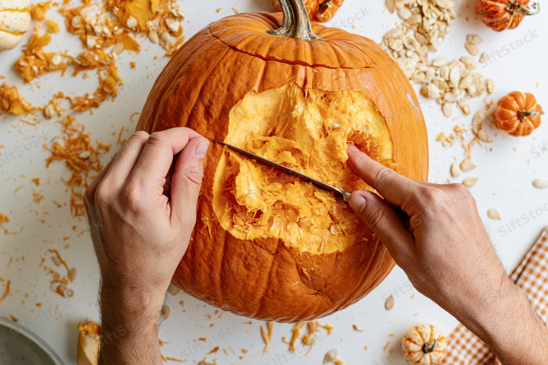Hands carving a pumpkin with a knife on a white surface surrounded by seeds and pulp