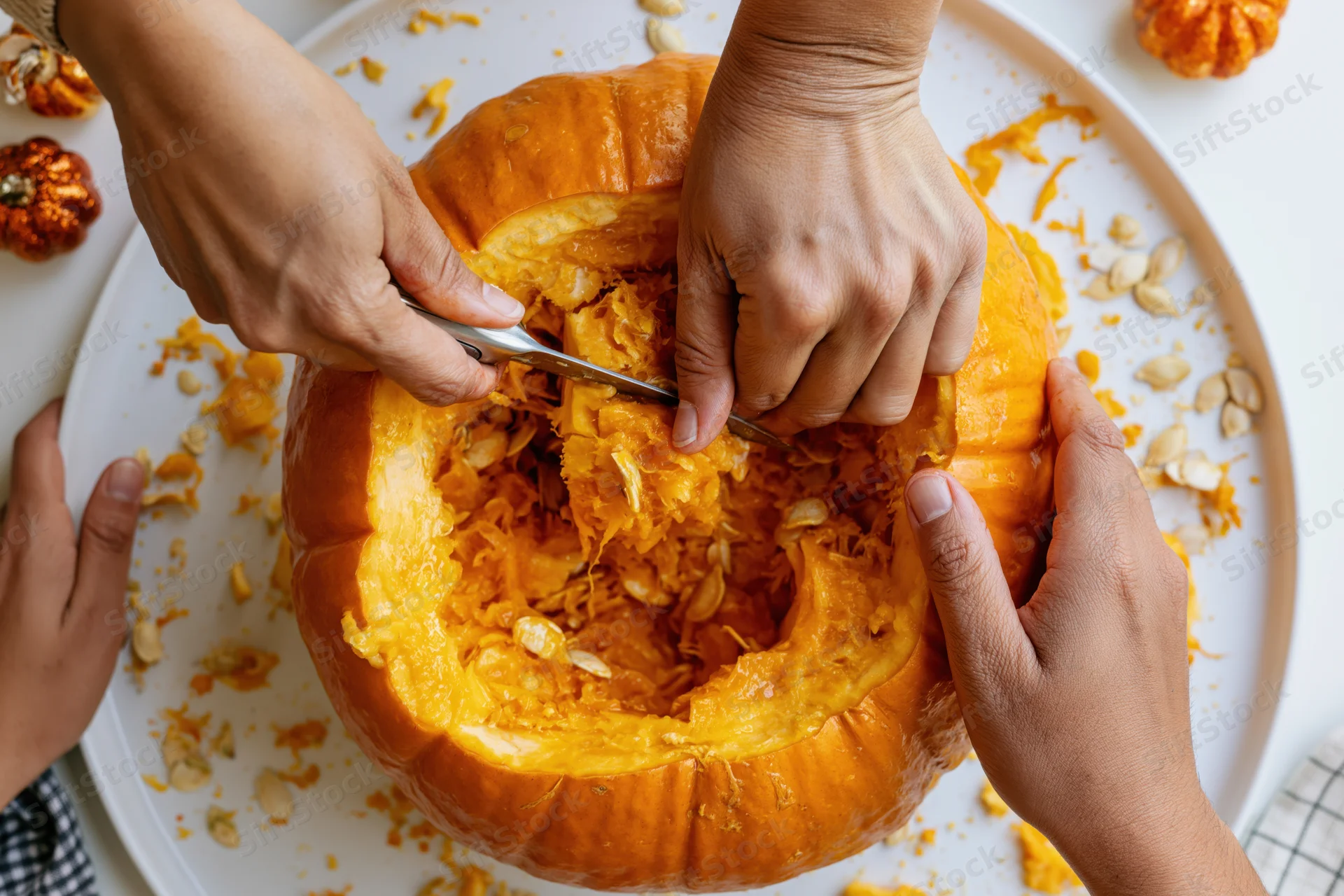 Hands carving a pumpkin on a white plate with seeds and pulp scattered around