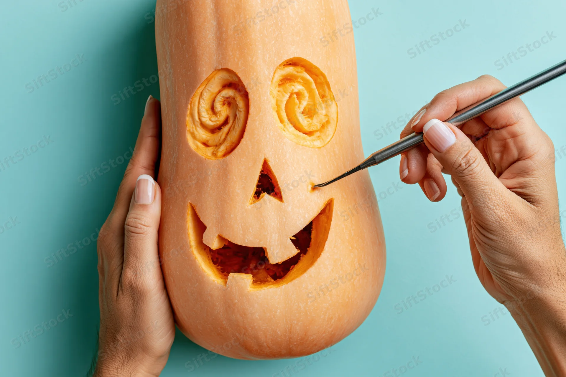 Hands carving a butternut squash jack-o-lantern with a paintbrush against a teal background