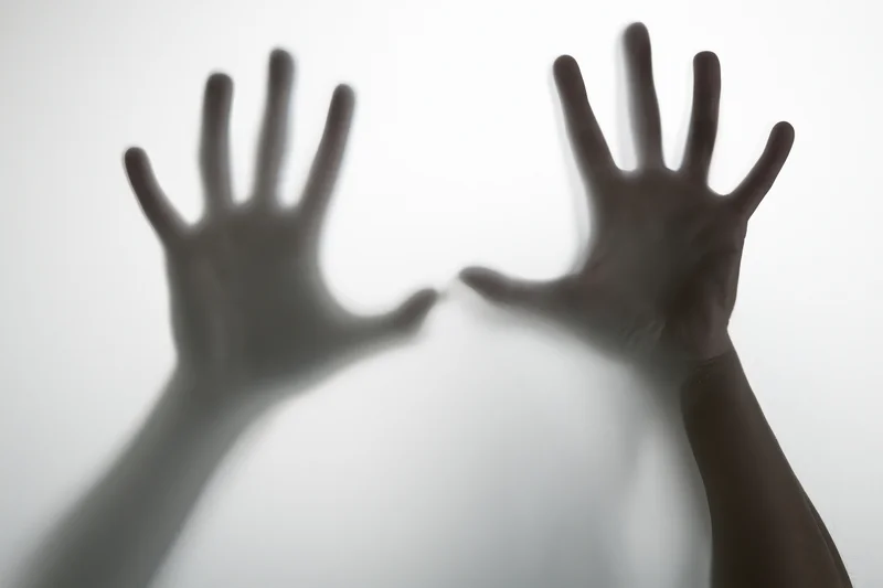 Two hands pressed against frosted glass with fingers spread, one hand in focus and the other blurred, creating a shadowy silhouette effect