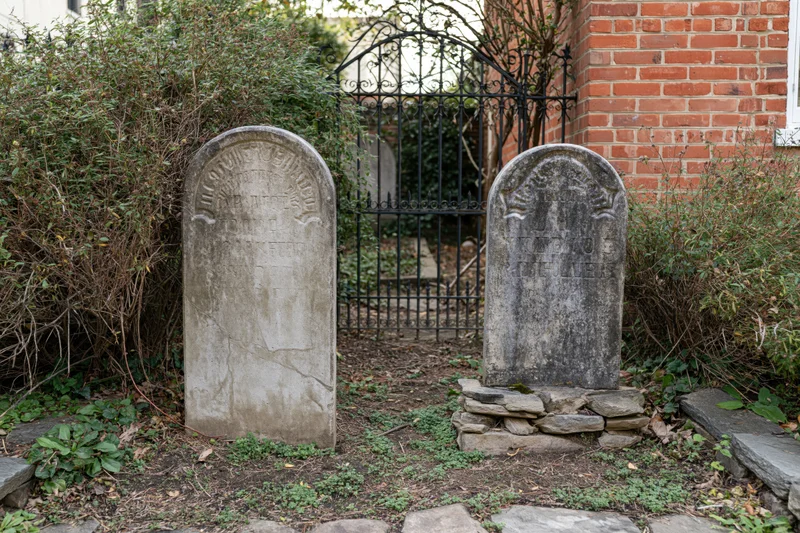 Two weathered gravestones with engraved epitaphs stand in a garden beside a brick wall and iron gate