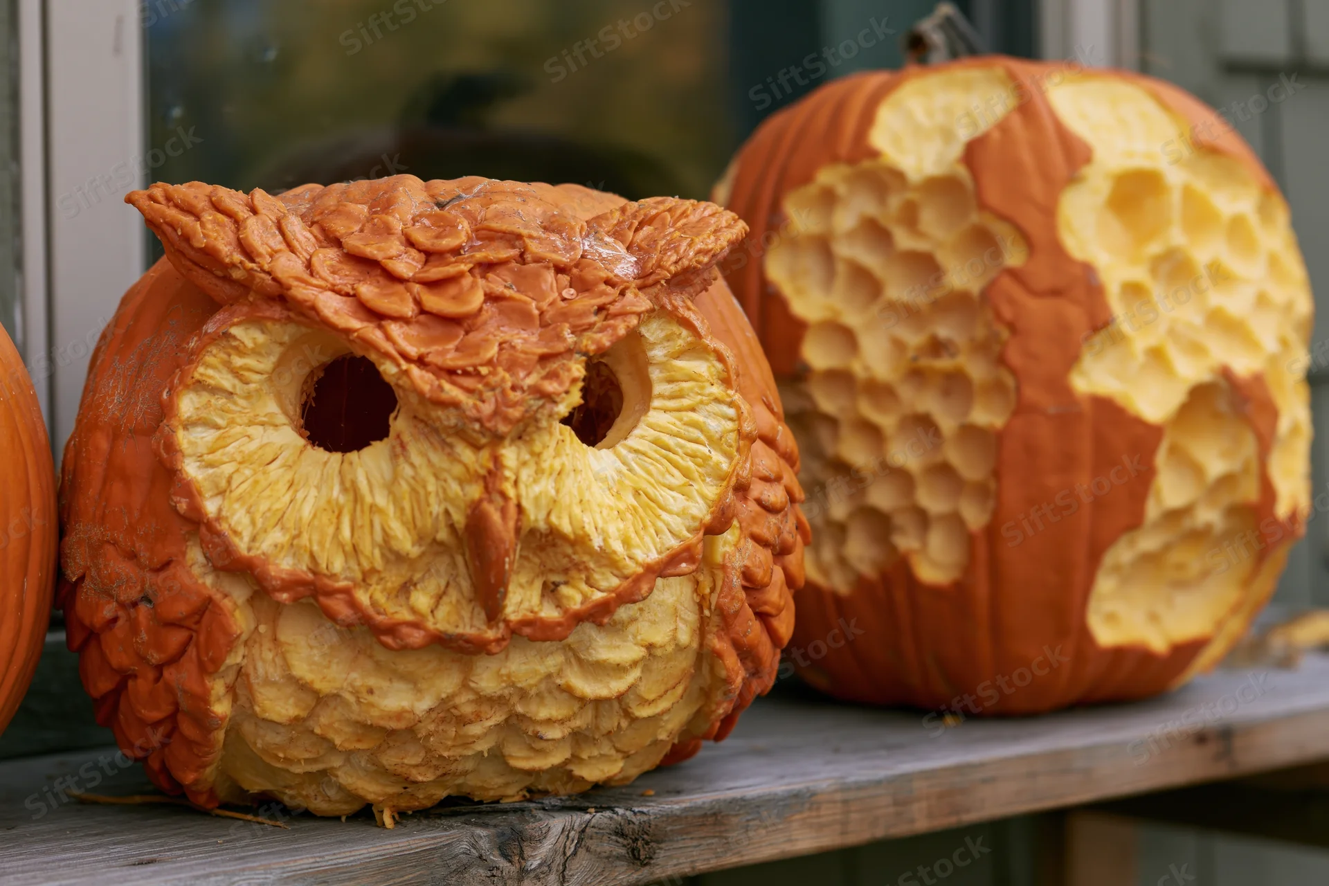 Intricately carved owl pumpkin and honeycomb-patterned pumpkin displayed on a wooden shelf outdoors near a house window