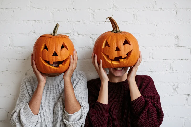 Two women holding carved pumpkins over their faces against a white brick wall
