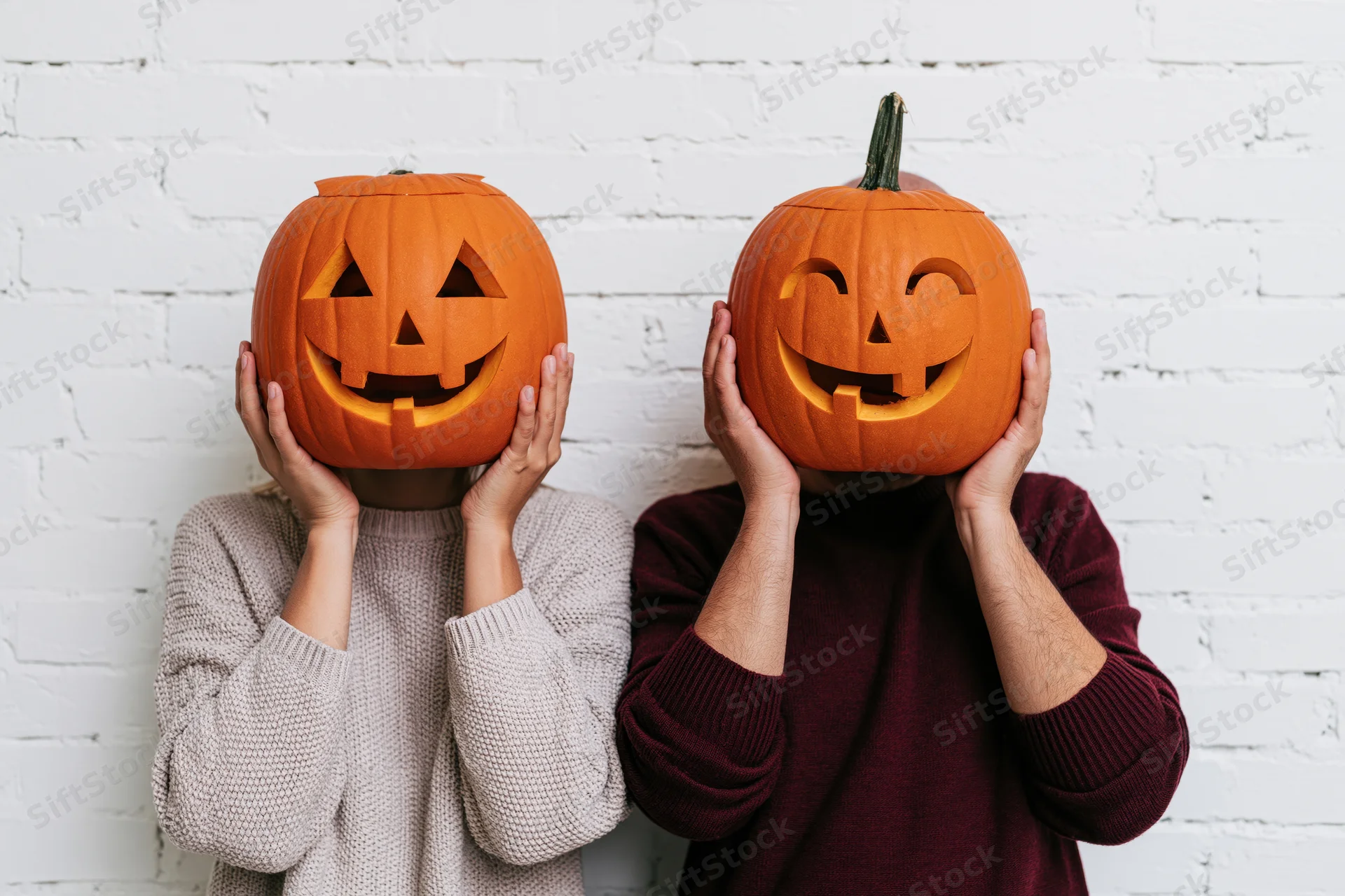 Two people holding carved pumpkins in front of their faces against a white brick wall