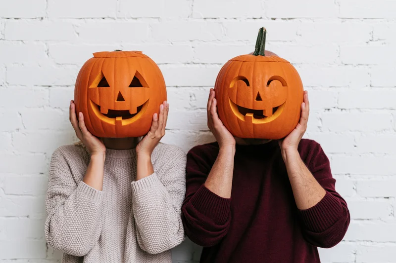 Two people holding carved pumpkins in front of their faces against a white brick wall