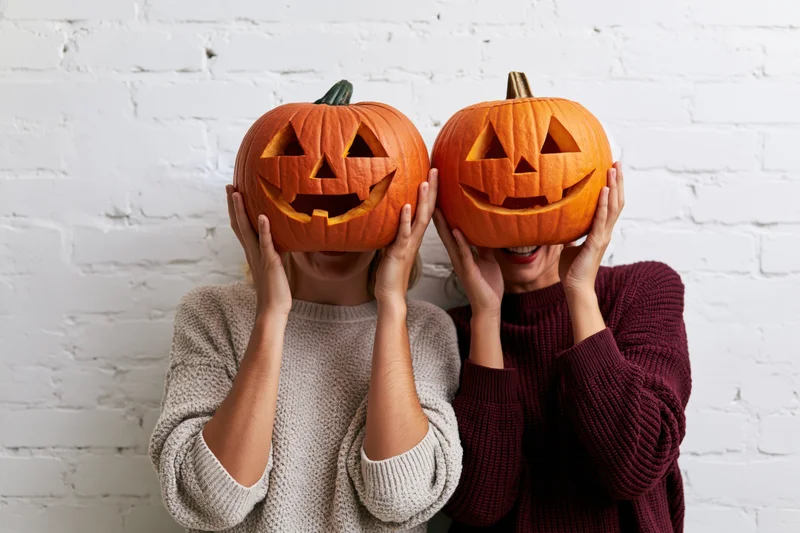 Two women holding carved jack-o-lanterns in front of their faces against a white brick wall