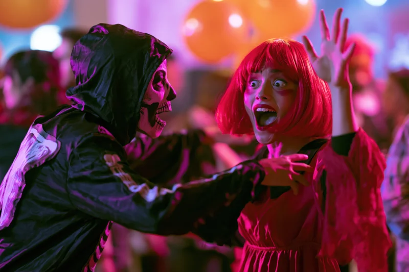 Woman in red wig reacting in surprise to man in skeleton costume at indoor halloween party