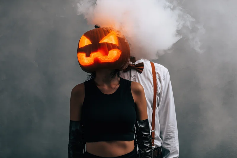 Woman with carved pumpkin head emitting smoke standing beside man in white shirt and suspenders against gray backdrop
