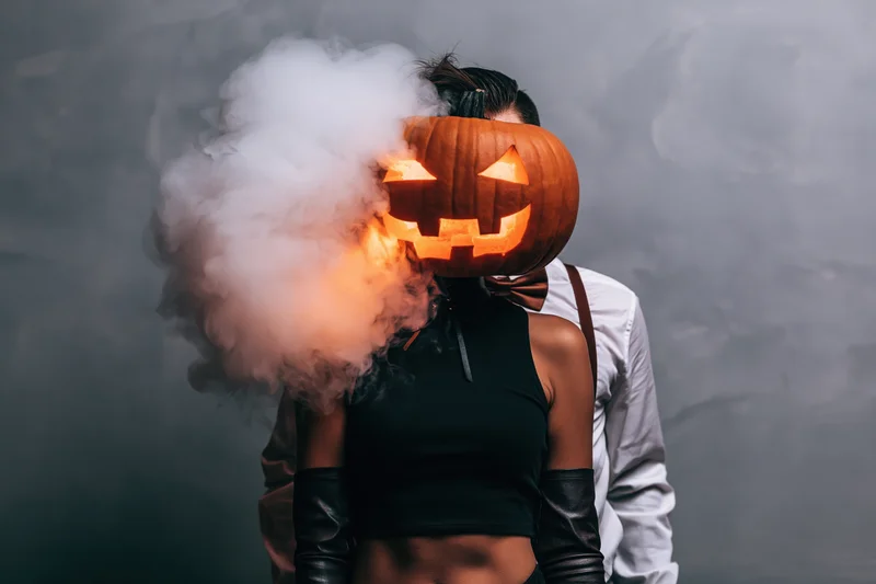Woman and man in halloween costumes with jack-o-lantern head and smoke effect in studio