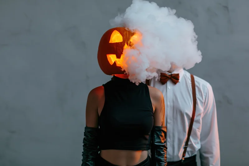 Woman with pumpkin head and man in bowtie and suspenders standing side by side in halloween costumes with smoke billowing around them against a gray wall