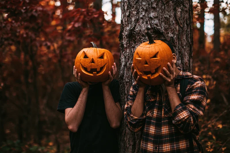Two men holding carved pumpkins over their faces beside a tree in an autumn forest