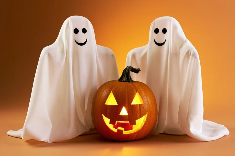 Two smiling ghost decorations flanking a glowing jack-o'-lantern on an orange background
