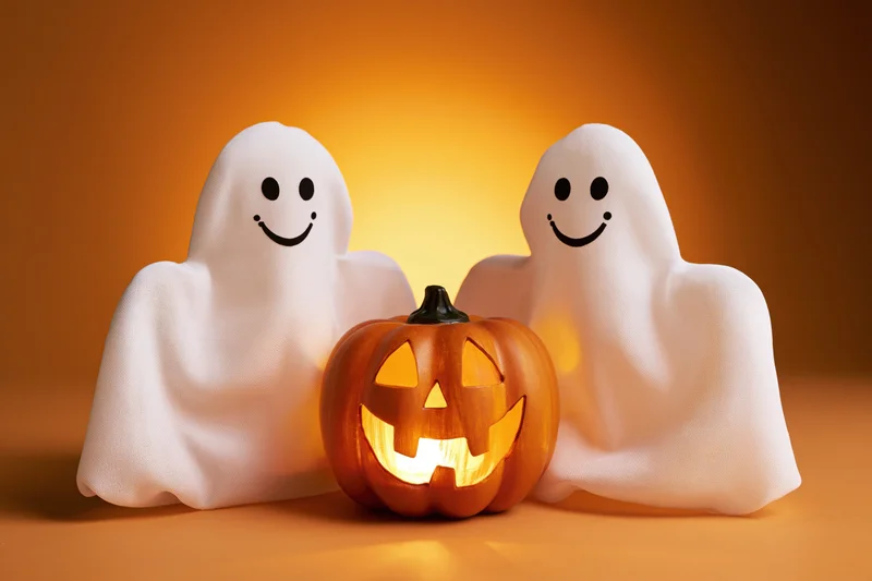 Two smiling ghost decorations flanking a glowing jack-o'-lantern on an orange surface