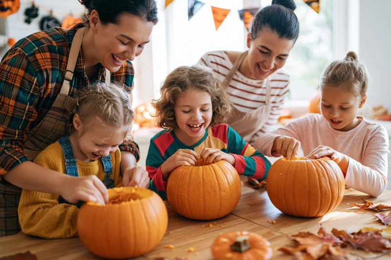 Two women and two children carving pumpkins together at a wooden kitchen table during halloween celebration