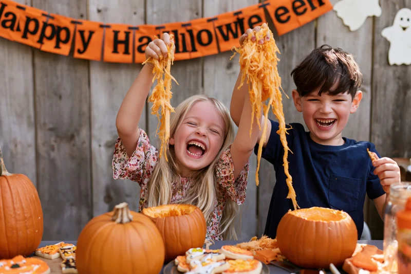 Two children laughing while scooping pumpkin guts at a halloween party outdoors