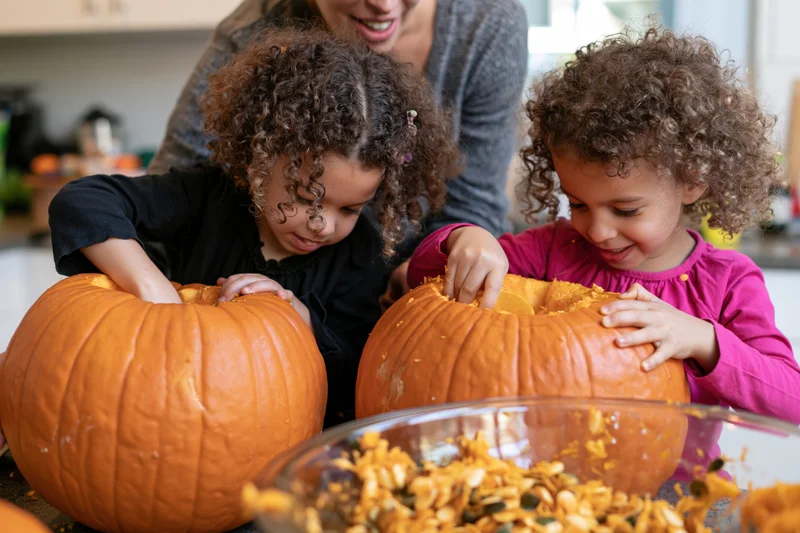 Two young girls scooping out pumpkins in a kitchen with their mother watching