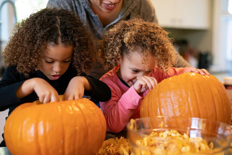 Two young curly-haired girls carving and scooping out pumpkins in a kitchen with their mother watching