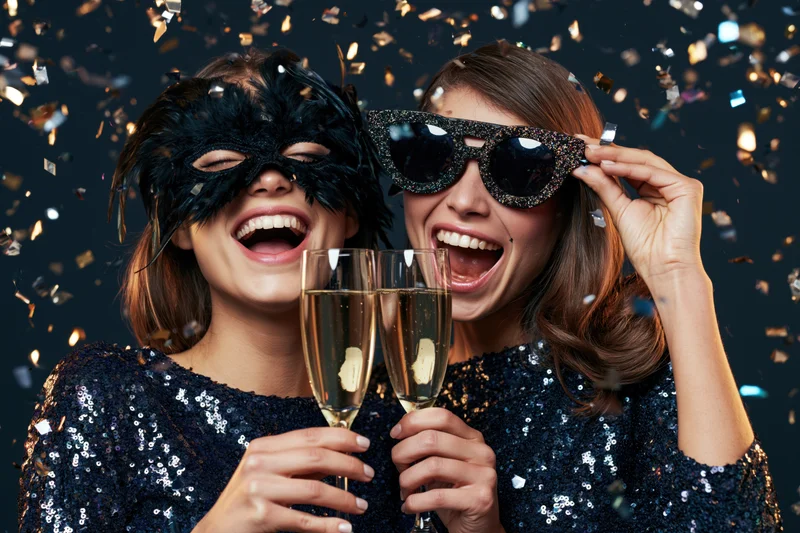 Two young women laughing and toasting with champagne flutes at a masquerade party with confetti falling around them