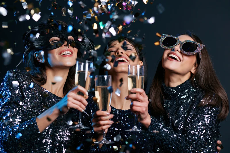 Three women in sequined dresses laughing and toasting with champagne glasses amidst falling confetti at a masquerade party