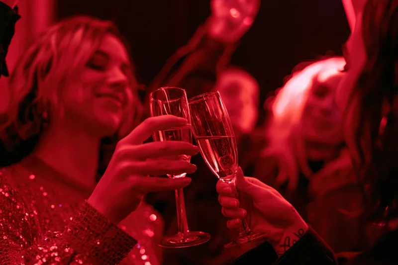 Two women clinking champagne flutes at a festive party under red lighting