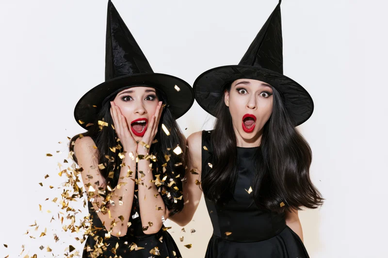 Two young women in witch costumes reacting with surprise amidst falling gold confetti in a studio setting