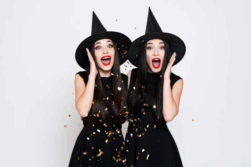 Two young women in witch costumes reacting with surprise amidst falling confetti in a studio setting