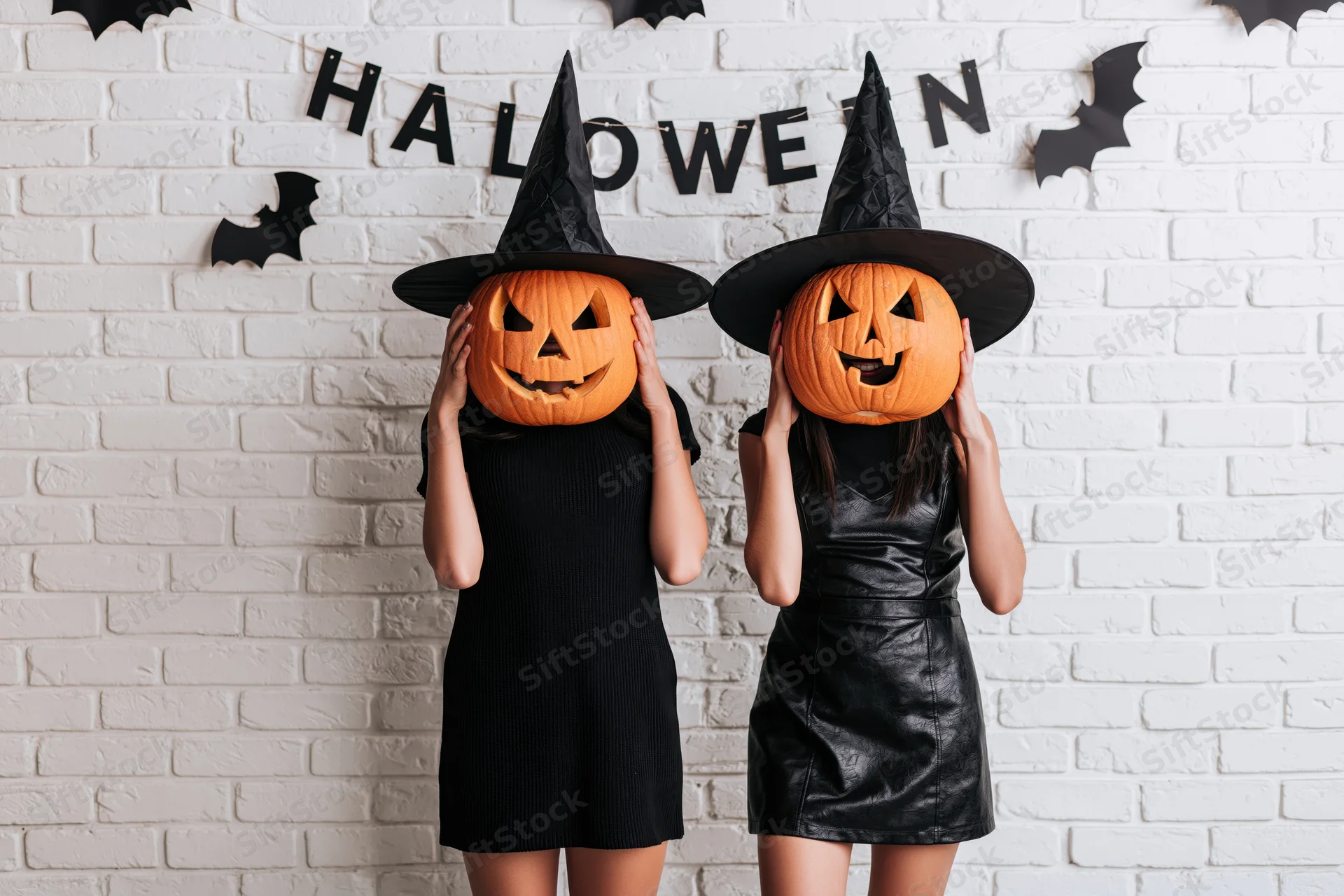Two women in witch hats holding jack-o-lanterns over their faces against a decorated brick wall