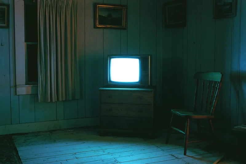 Vintage crt television glowing blue-white on wooden dresser in dark room with chair and framed pictures on wood-paneled walls