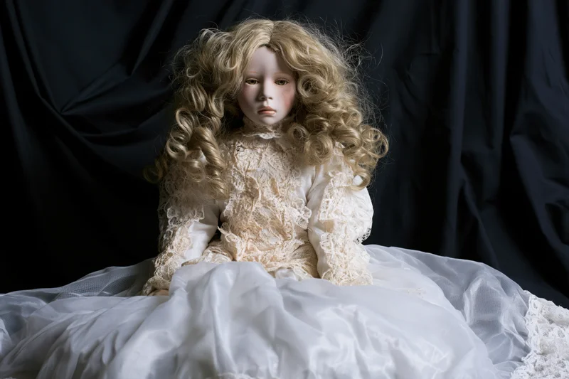 Porcelain doll with curly blonde hair seated in white lace dress against dark backdrop