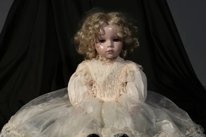 Vintage porcelain doll with curly blonde hair seated in a white lace dress against a black backdrop