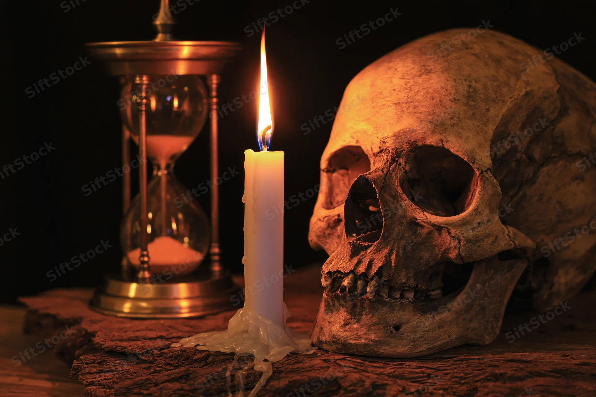 Weathered human skull beside lit candle and hourglass on wooden surface