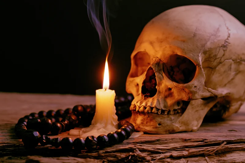 Weathered human skull beside a lit candle with smoke and a dark rosary on rough wood