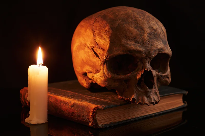 Weathered human skull resting on an old worn book beside a lit candle on a reflective surface