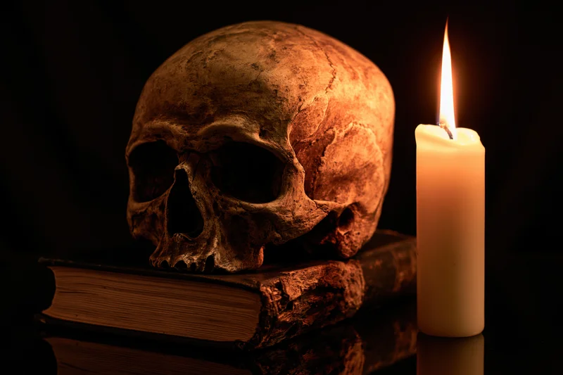 Weathered human skull resting on an old worn book beside a lit candle in dark setting