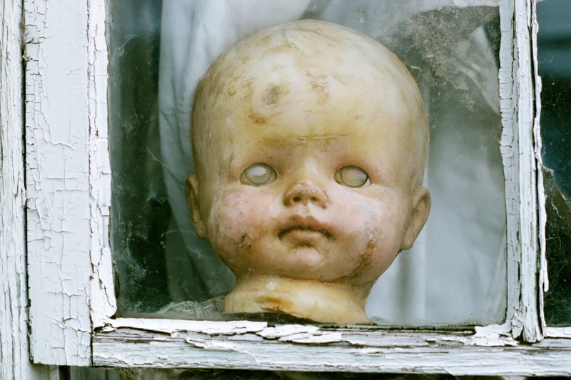 Weathered plastic doll head pressed against glass in a peeling white window frame