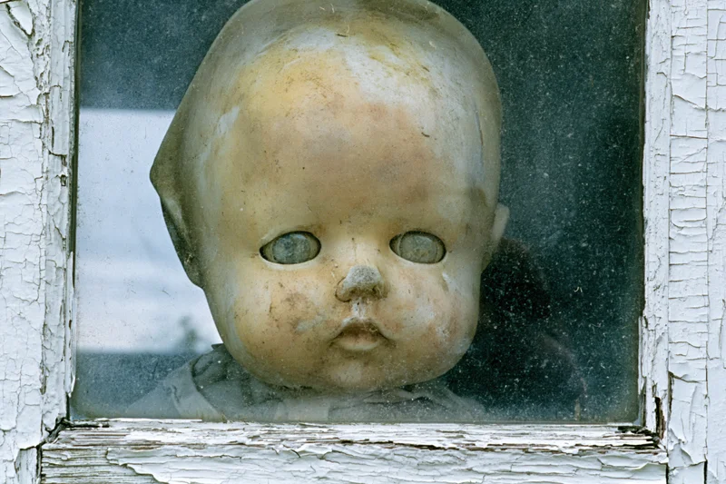 Weathered plastic doll head pressed against glass in a peeling white window frame