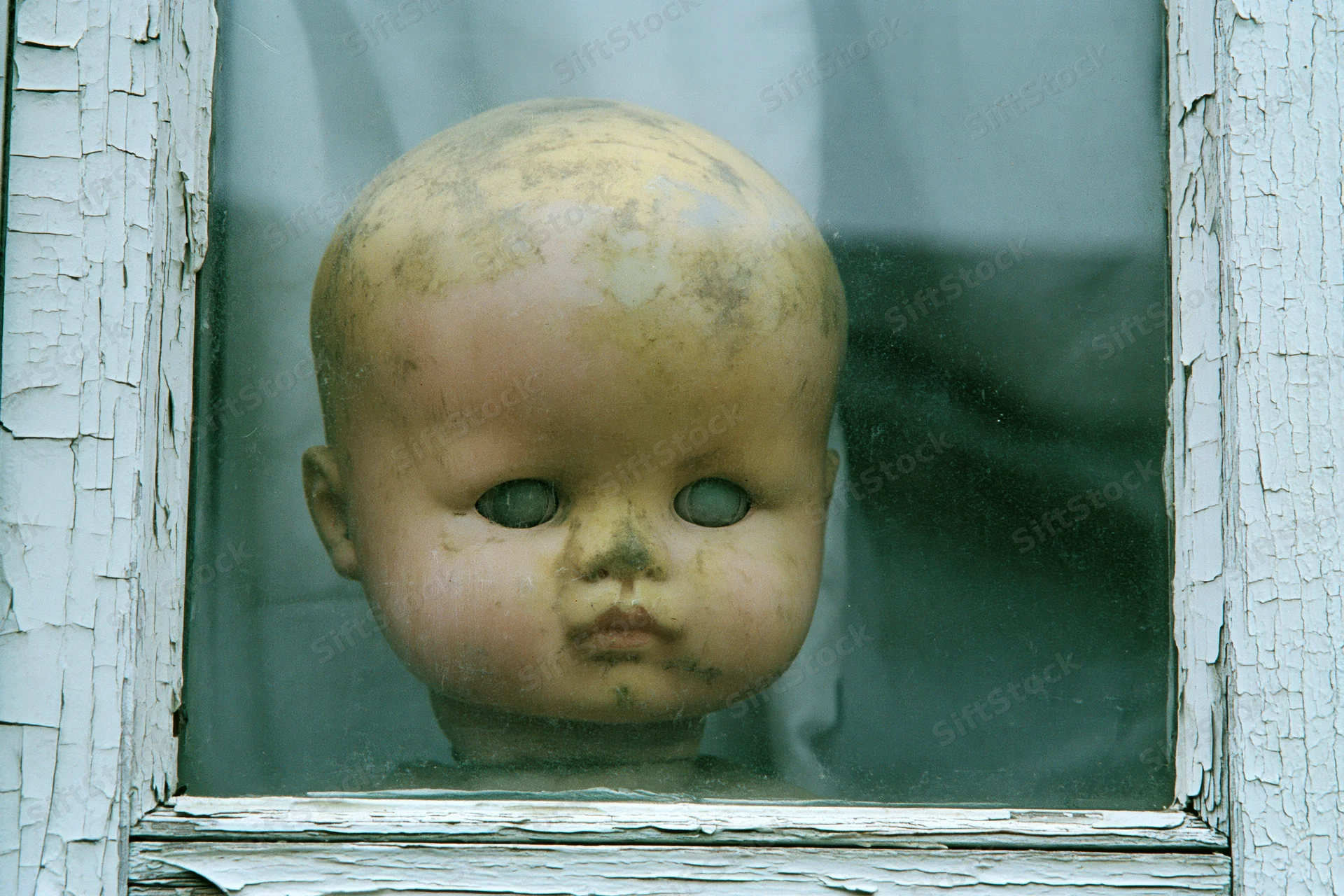 Weathered plastic doll head pressed against glass window in peeling white frame