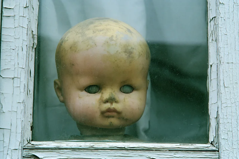 Weathered plastic doll head pressed against glass window in peeling white frame