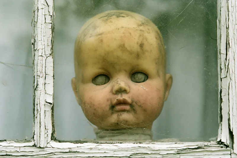 Weathered plastic doll head pressed against a dirty windowpane in a dilapidated house