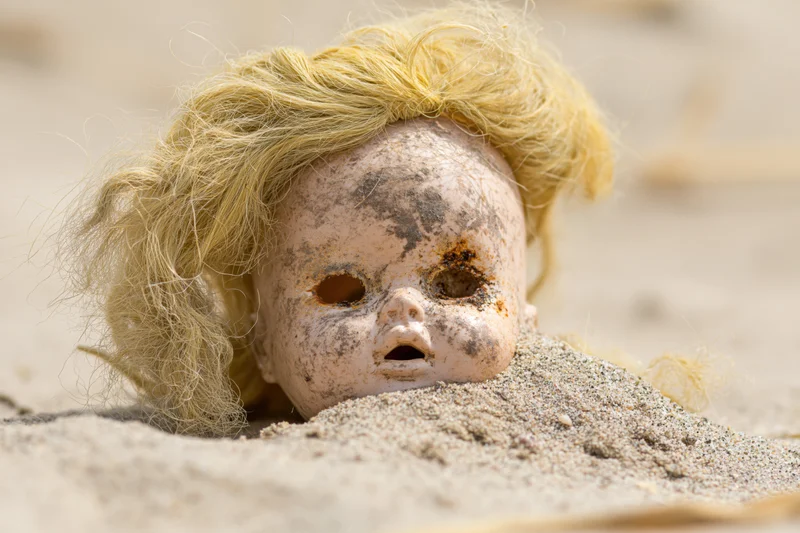 Weathered plastic doll head half-buried in dry sand with blonde hair and empty eye sockets