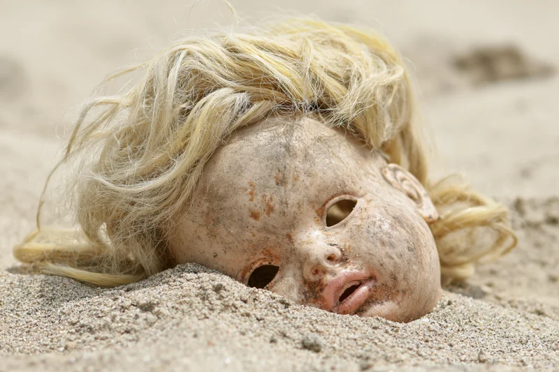 Weathered plastic doll head half-buried in dry sand with open mouth and blonde hair strands visible