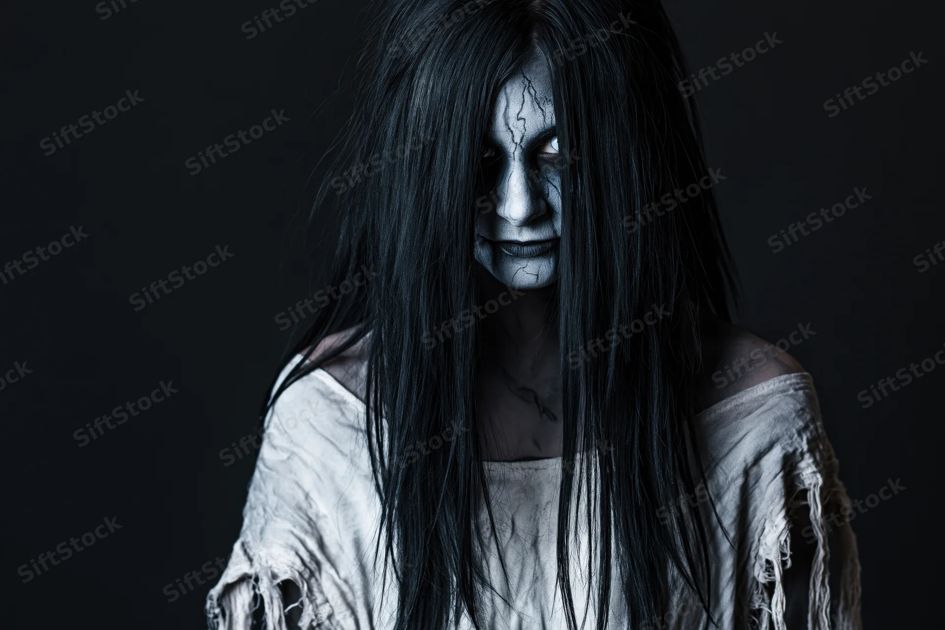 Woman in ghost makeup with cracked white face paint and long black hair, wearing tattered off-shoulder dress, studio portrait